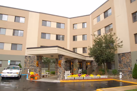 Exterior view of a multi-story senior living facility with a covered entrance supported by stone pillars. The building has beige and light brown walls with several windows. There are autumn decorations including pumpkins and flowers near the entrance, and a parked car is visible on the left side.