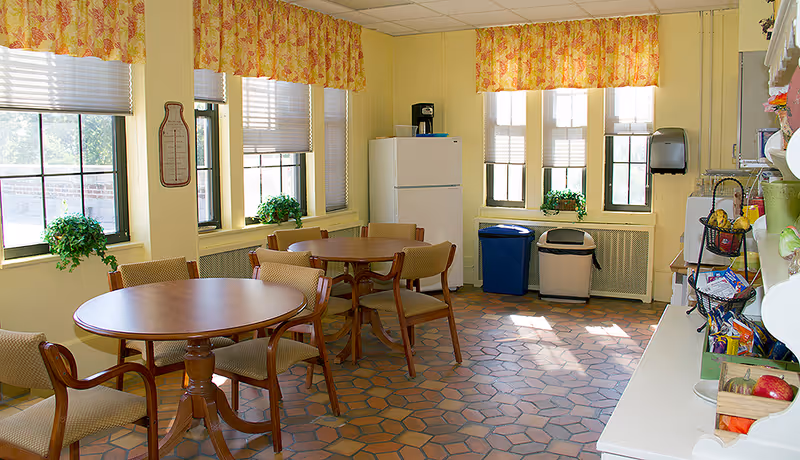 A bright room with yellow walls and large windows covered with floral valances. The room contains two round wooden tables surrounded by cushioned chairs. There is a white refrigerator in the corner, a coffee maker on top of it, and two trash bins near the windows. On the right side, there is a counter with various snacks and a microwave. The floor is tiled with hexagonal terracotta tiles.