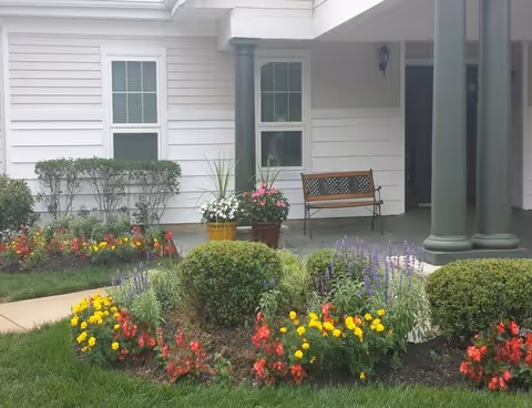 A garden area with colorful flowers and green bushes in front of a white building with two windows and a covered porch. The porch has two large green columns and a wooden bench.