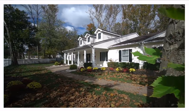 A white single-story building with a porch and columns, surrounded by a garden with colorful flowers and trees with autumn leaves. A paved walkway leads to the entrance, and a white picket fence is visible in the background under a partly cloudy sky.