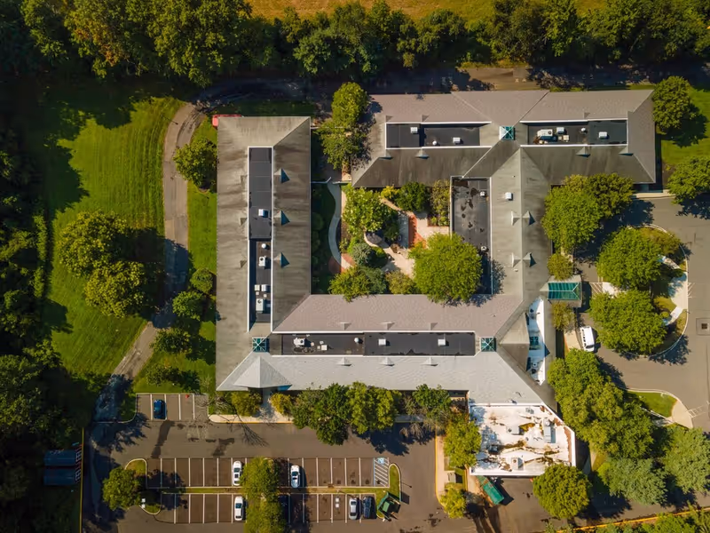 Aerial view of a senior living facility named Complete Care at Marcella, showing the building's roof, surrounding trees, parking lot with several cars, and landscaped green areas.