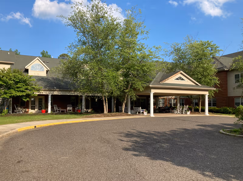 Exterior view of Woodview Estates facility showing a covered entrance with white columns, surrounded by trees and greenery under a blue sky with some clouds. The building has a brick and siding facade with multiple windows and a paved driveway in front.