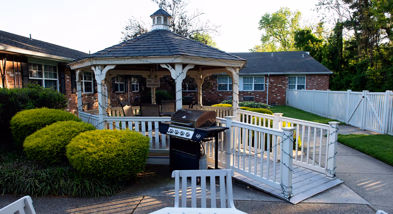 Outdoor patio area at Wynwood Rehabilitation and Healthcare Center featuring a white gazebo with seating inside, a black barbecue grill in front, surrounded by green bushes and a white fence. The background shows a brick building and trees.