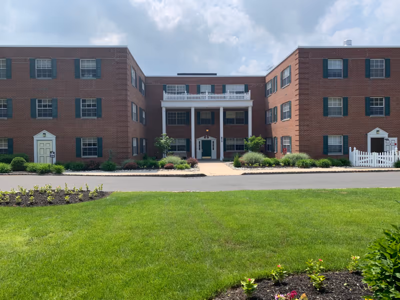Front view of a three-story red brick building with green shutters, a central columned entrance, landscaped beds and a lawn in the foreground.