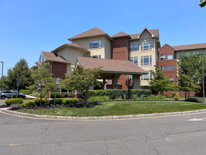 Exterior view of a multi-story senior living facility building with a covered entrance, surrounded by well-maintained landscaping including trees, bushes, and flower beds under a clear blue sky.