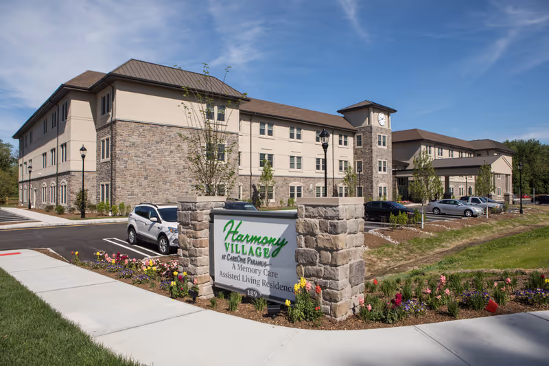 Exterior front view of Harmony Village at CareOne Paramus, a three-story senior living building with a stone entrance sign and landscaped grounds.