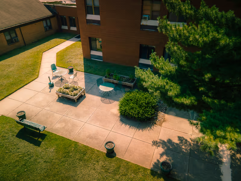 Outdoor patio area at Complete Care at Burlington Woods with concrete walkways, a few chairs and tables, planters with flowers and greenery, a bench, and a large tree casting shadows on the ground.