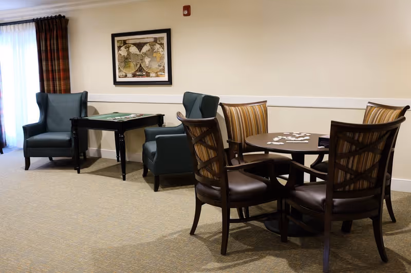 A cozy seating area in a senior living facility with two blue armchairs next to a small table with a game on it, and a round table with four striped cushioned chairs around it. There is a framed world map on the wall and plaid curtains covering a window or door.