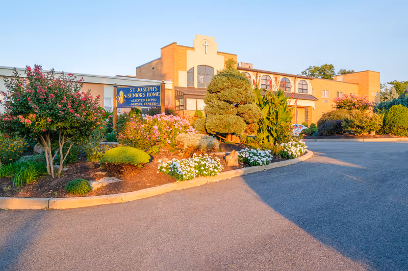 Exterior view of St. Joseph's Seniors Home, an assisted living and nursing center. The building is a two-story brick structure with a cross symbol above the entrance. In front of the building, there is a well-maintained garden with various shrubs, flowering plants, and a sign displaying the facility's name. The sky is clear and blue.