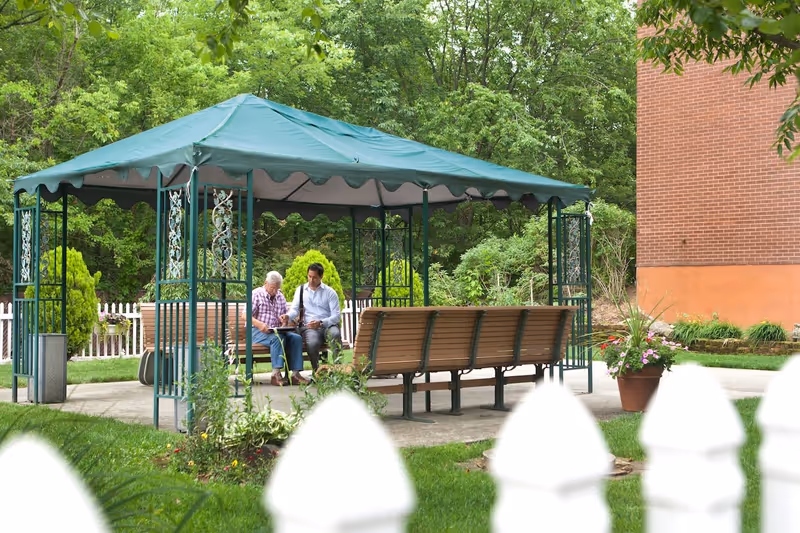 Two people sitting and talking under a green canopy gazebo in a garden area with trees, plants, and a white picket fence. There is a brick building visible on the right side and a flower pot with pink flowers near the gazebo.