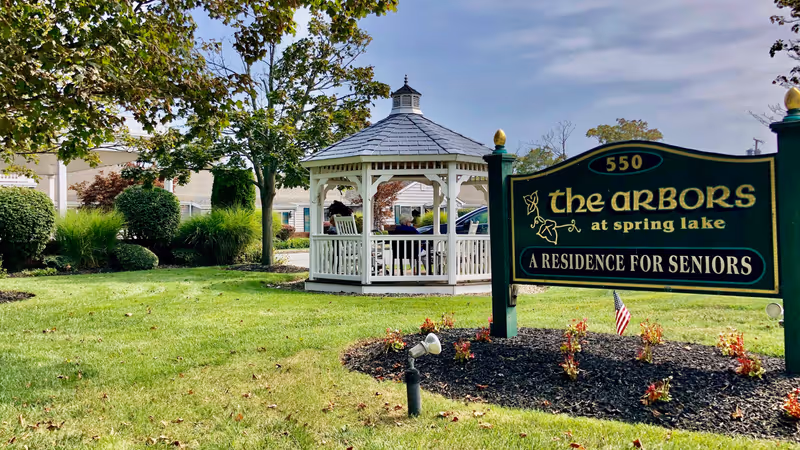 Outdoor view of The Arbors at Spring Lake senior living facility showing a green lawn with a white gazebo in the background and a sign in the foreground that reads '550 the arbors at spring lake A RESIDENCE FOR SENIORS'.