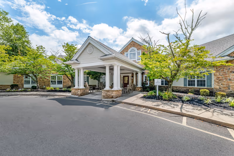 Front entrance porte-cochere of a senior living building with white columns, outdoor seating, and landscaped trees under a partly cloudy sky.