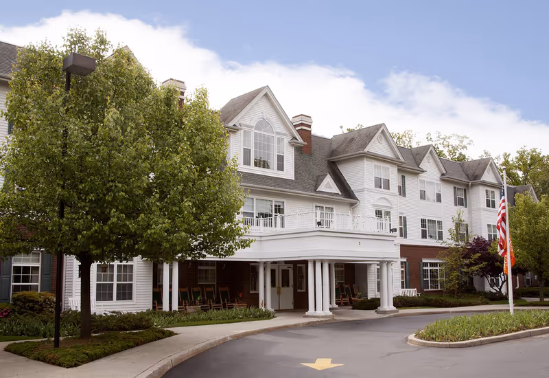 Exterior view of a multi-story senior living facility with white siding and brick accents. The building features large windows, a covered entrance with white columns, rocking chairs on the porch, a curved driveway, and landscaped greenery including trees and shrubs. An American flag is displayed on a flagpole near the entrance.