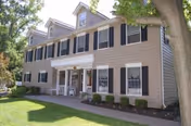 Exterior view of a two-story beige building with black shutters and white trim, featuring a covered entrance with white columns. The building is surrounded by a well-maintained lawn, shrubs, and trees under a clear sky.