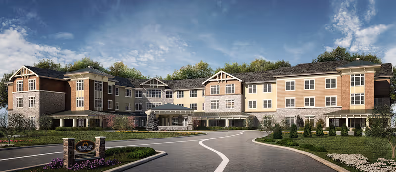 Exterior view of a large three-story senior living facility building with a curved driveway leading to the entrance. The building features a mix of stone and siding with multiple windows and a covered entrance. There is a sign in front that reads 'Sunrise of Bridgewater' surrounded by landscaping and greenery under a partly cloudy sky.