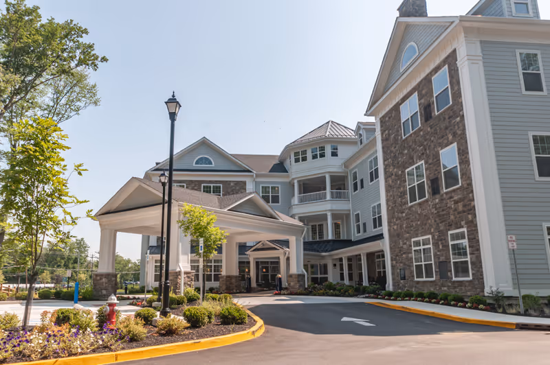 Exterior view of Arbor Terrace Marlton, a multi-story senior living facility with a covered entrance, stone and siding facade, landscaped garden beds, and a clear blue sky.