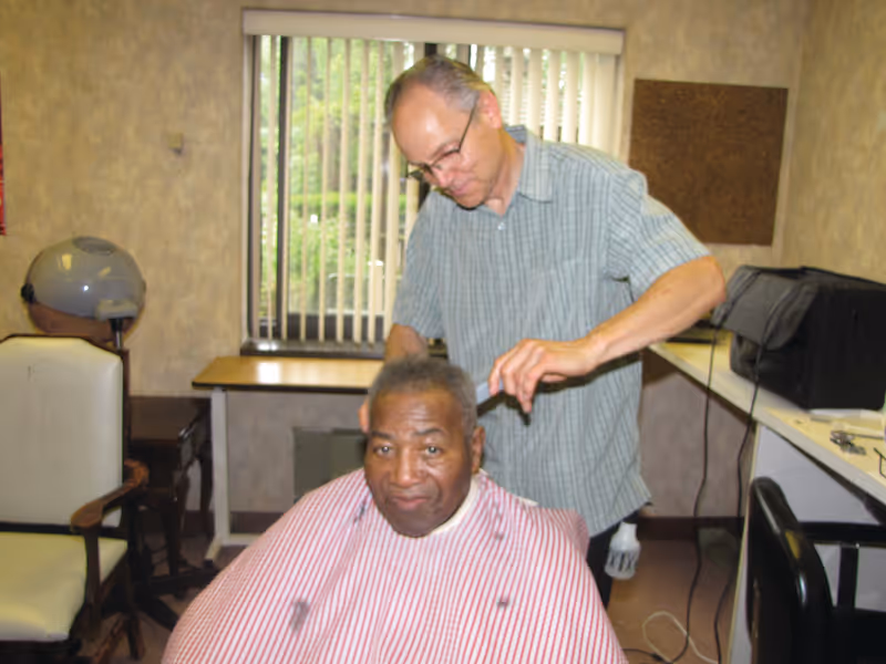 A man is getting a haircut from a barber in a small room with a window that has vertical blinds. The man receiving the haircut is wearing a red and white striped barber cape and looking at the camera. The barber is standing behind him, focused on cutting his hair. The room has a chair, a hair dryer, and a counter with various items on it.