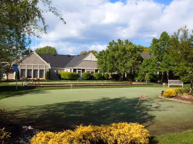 A putting green with small red flags surrounded by landscaped bushes and trees in front of a single-story building under a partly cloudy sky.