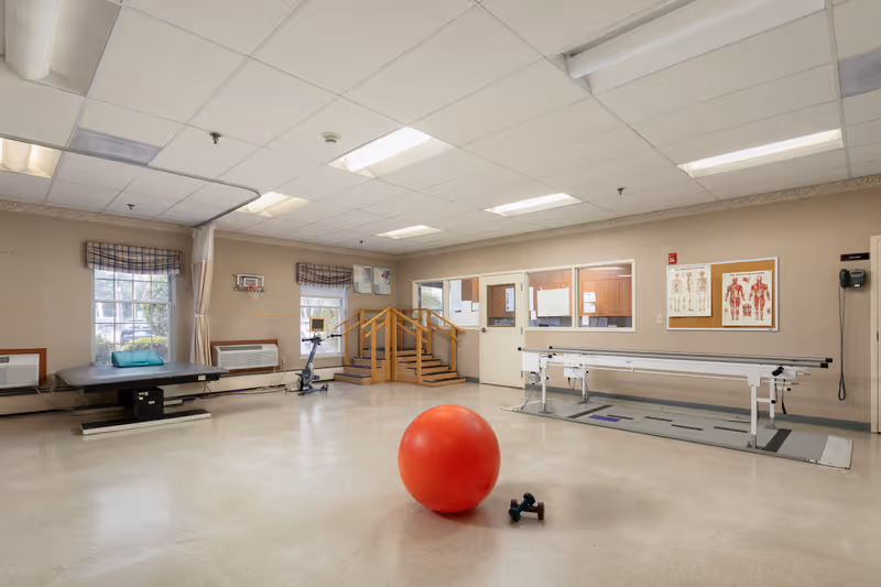 A spacious physical therapy room with a large red exercise ball and two small dumbbells on the floor. The room features a therapy table, an exercise bike, a set of wooden stairs with handrails for rehabilitation, and parallel bars for walking exercises. Anatomical charts are displayed on the wall, and windows with plaid valances allow natural light to enter.