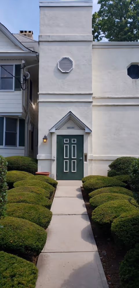 A concrete pathway lined with neatly trimmed green bushes leads to a green door with white trim on a light-colored building. The building has a small peaked roof above the door, a wall-mounted lantern light to the left of the door, and a vent above the door. Part of an adjacent white building with windows is visible on the left side, and trees can be seen in the background.