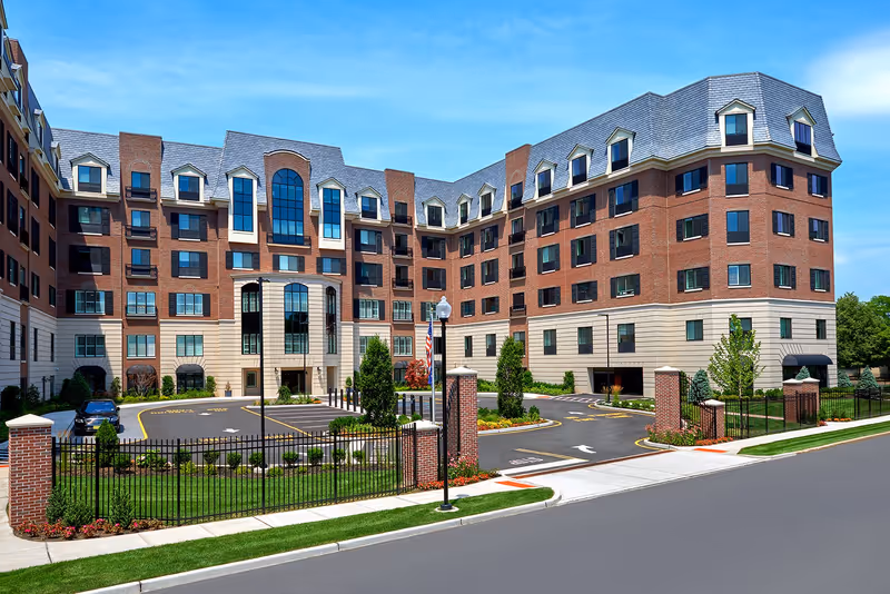 Exterior view of The Bristal Assisted Living at Englewood, a large multi-story brick building with numerous windows, a gated entrance, landscaped greenery, and a clear blue sky.