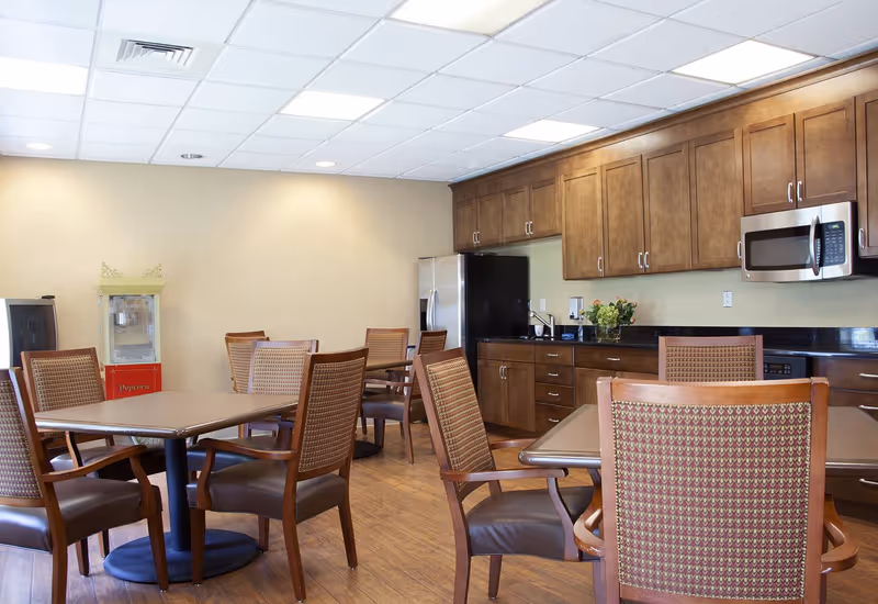 A dining area with several wooden tables and chairs with cushioned seats and backs. The room has wooden flooring and a kitchen area with wooden cabinets, a stainless steel refrigerator, microwave, and a black countertop. There is a popcorn machine in the corner and a vase with flowers on the counter.