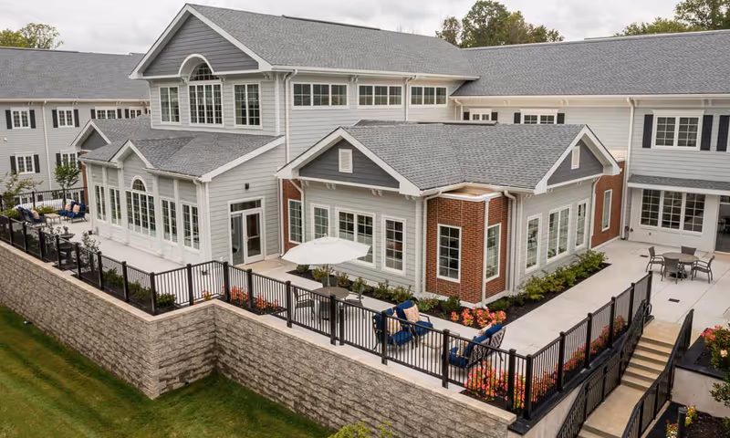 Exterior view of a senior living facility building with gray siding and brick accents, featuring multiple large windows and a fenced patio area with outdoor seating and tables with umbrellas. The building is surrounded by a stone retaining wall and landscaped with flowers and shrubs.
