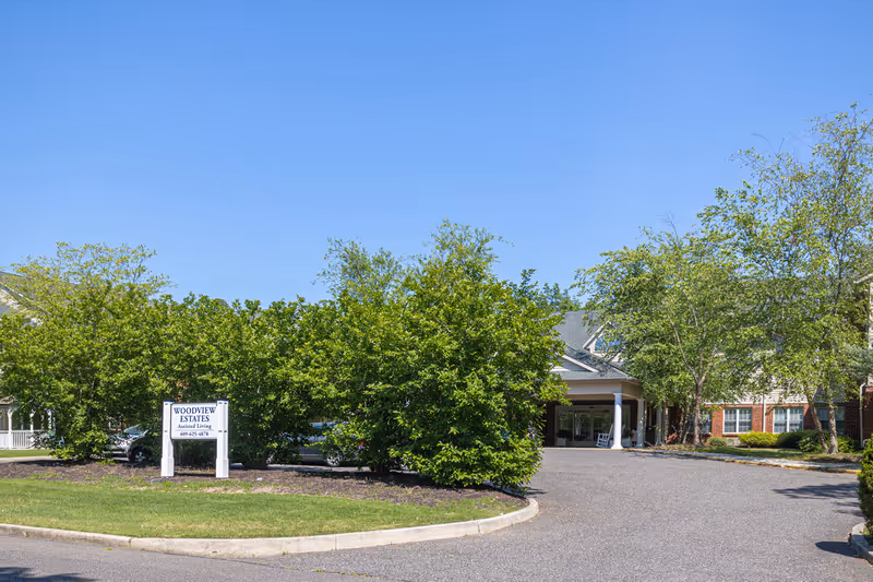 Exterior view of Woodview Estates assisted living facility showing the driveway entrance, green bushes, trees, and part of the building under a clear blue sky.