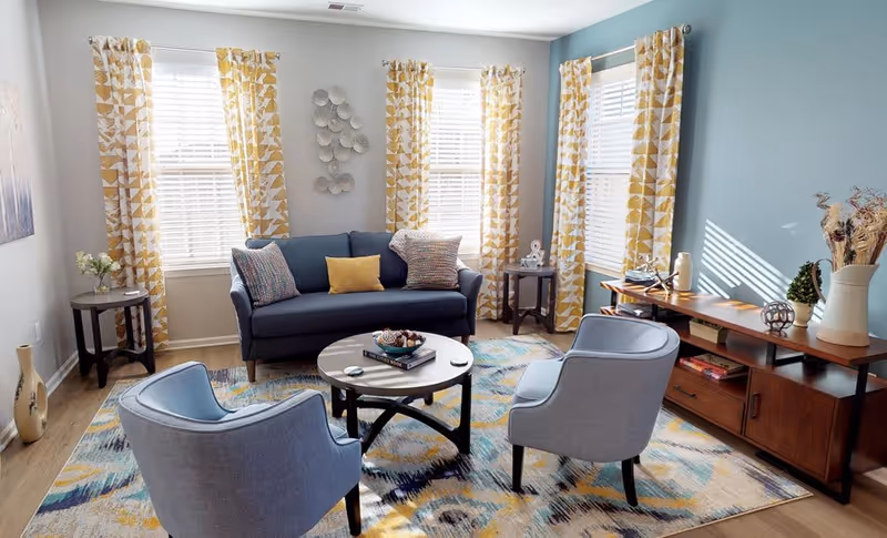 A bright and cozy living room with a blue sofa adorned with colorful cushions, two light blue armchairs, a round wooden coffee table with books and decorative items, and a patterned area rug. The room has three windows with yellow and white patterned curtains, light blue and white walls, and a wooden sideboard with decorative vases and plants.