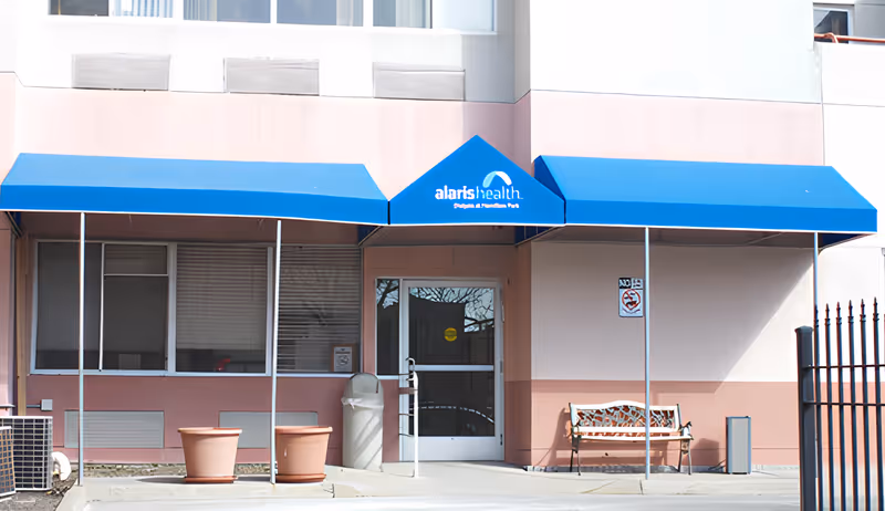 Entrance to Alaris Health at Cedar Grove with a blue awning above the door displaying the facility's name. There are two large empty plant pots, a trash can, a bench, and a no smoking sign near the entrance.