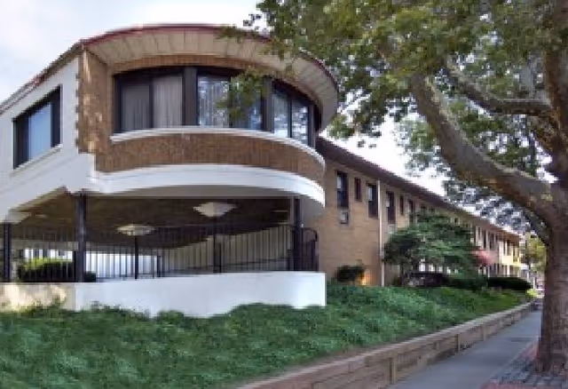 Exterior view of a two-story brick building with large windows and a covered balcony area, surrounded by greenery and a tree near the sidewalk.