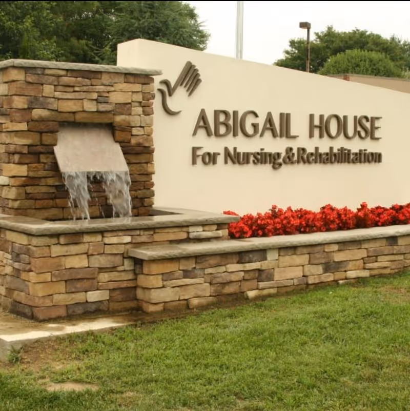Stone fountain and landscaped entrance sign reading 'ABIGAIL HOUSE For Nursing & Rehabilitation' with red flowers and grass.