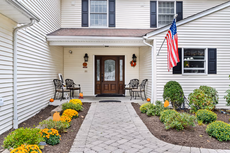 Covered front entrance of a light-colored building with a paved walkway, flower beds, seating, and an American flag.