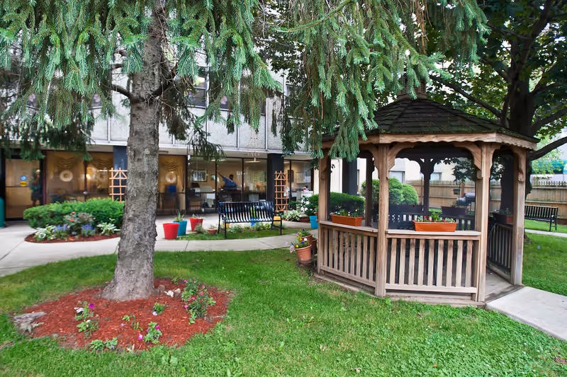 Outdoor garden area at Windsor Gardens Care Center featuring a wooden gazebo with flower pots, green grass, a large tree with hanging branches, benches, and a building with large windows in the background.