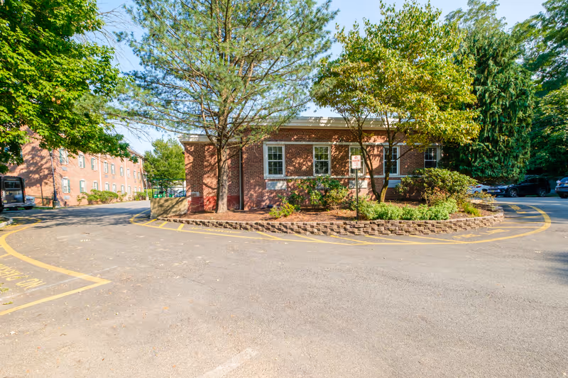 Brick single-story building surrounded by trees with a circular driveway and parking area in front.