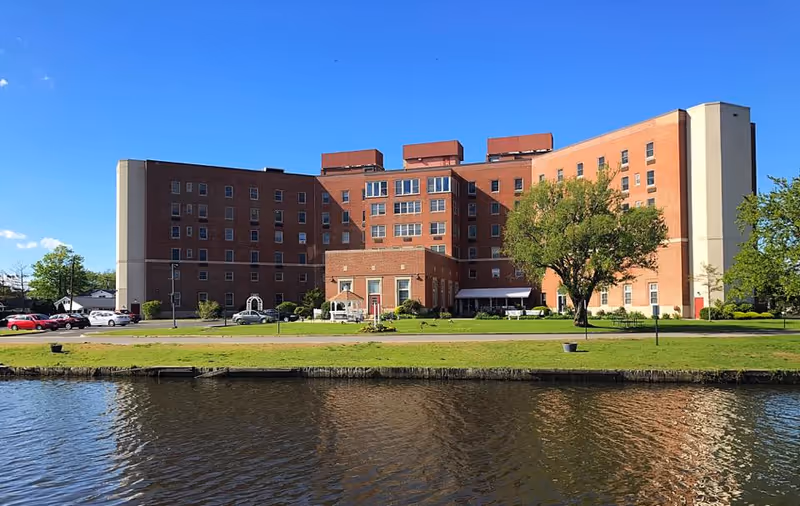 Front view of a large brick multi-story senior living building beside a river with a lawn and trees under a clear blue sky.