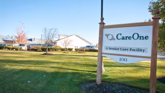 A lawn and wooden sign reading "CareOne - A Senior Care Facility - 100" with the facility building visible in the background.