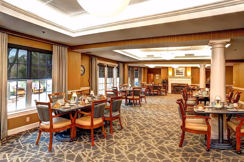 A spacious dining room with multiple wooden tables and chairs arranged neatly. Each table is set with plates, cups, napkins, and silverware. Large windows with curtains allow natural light to enter the room. The room features a patterned carpet, warm-toned walls, a fireplace at the far end, and ceiling lights providing bright illumination.