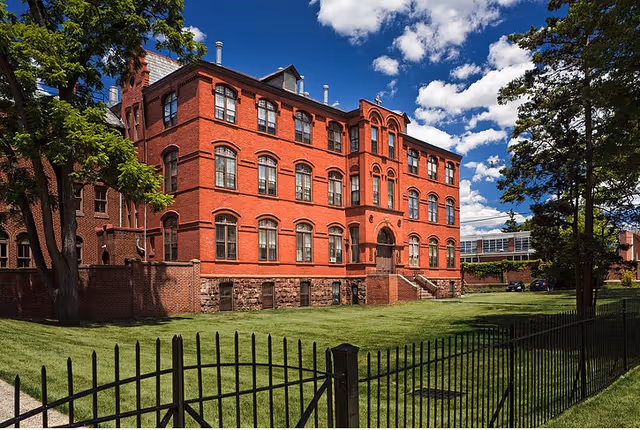 Red brick three-story historic building with arched windows and a fenced green lawn under a blue sky.