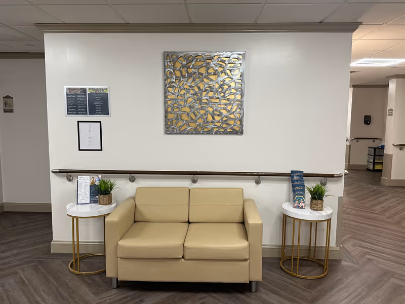 A beige loveseat sofa is centered against a white wall with a decorative metal wall art above it. On each side of the sofa, there are small round tables with white marble tops and gold legs, each holding a small potted plant and informational brochures. The floor has a wood-patterned design, and there are handrails along the wall. The hallway extends on both sides with doors and signage visible.