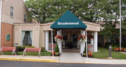 Entrance to a building with a green canopy labeled 'Brendenwood'. The building has beige walls and large windows with white curtains. There are two red benches on the left side near the entrance, and flower pots with red and white flowers hanging on white pillars. Trees and greenery surround the area.