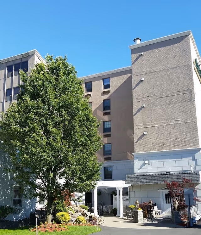 Exterior view of a multi-story senior living facility building with a large green tree and landscaped garden area in front. There is a covered entrance with white columns and outdoor seating visible near the entrance under a clear blue sky.