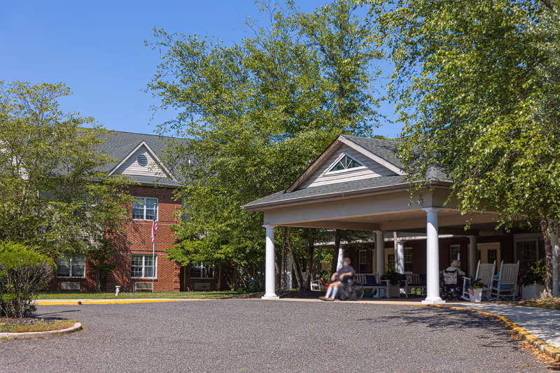 Exterior view of Woodview Estates senior living facility showing a brick building partially obscured by trees and a covered entrance with white columns. Two people in wheelchairs are near the entrance, and there are rocking chairs on the porch. The sky is clear and blue.