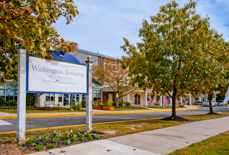 Exterior view of Washington Township Senior Living facility with a large sign in the foreground. The building is surrounded by trees with autumn foliage, a sidewalk, and a driveway. American flags are displayed near the entrance.