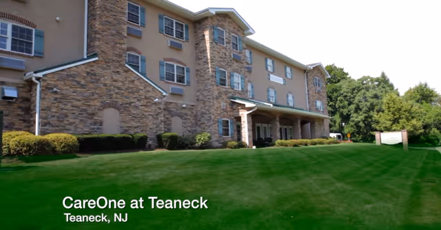 Exterior view of CareOne at Teaneck, a multi-story senior living facility with a stone and brick facade, surrounded by green grass and trees under a clear sky.