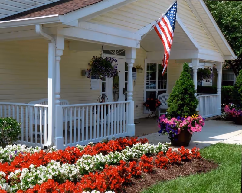 Front porch of a yellow building with white railings and columns, decorated with hanging flower baskets and potted plants. An American flag is displayed on the porch. In front of the porch is a garden bed with red and white flowers and a neatly trimmed green lawn.