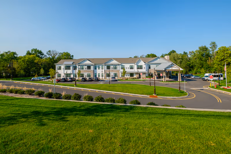 Exterior view of Arbor Terrace Basking Ridge, a two-story senior living facility with a large parking area, green lawns, and trees surrounding the building under a clear blue sky.