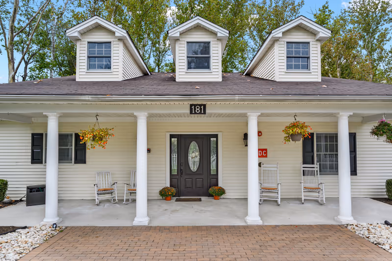 Front entrance of a light-colored building with a covered porch, white columns, rocking chairs, hanging plants, and the number 181 above the door.