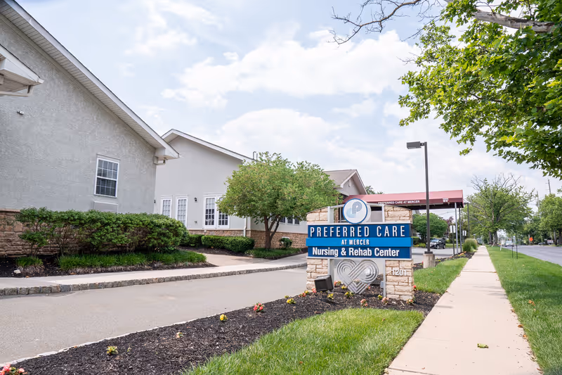Exterior view of Preferred Care at Mercer Nursing & Rehab Center showing the building, a sign with the facility name, a sidewalk, trees, and a partly cloudy sky.