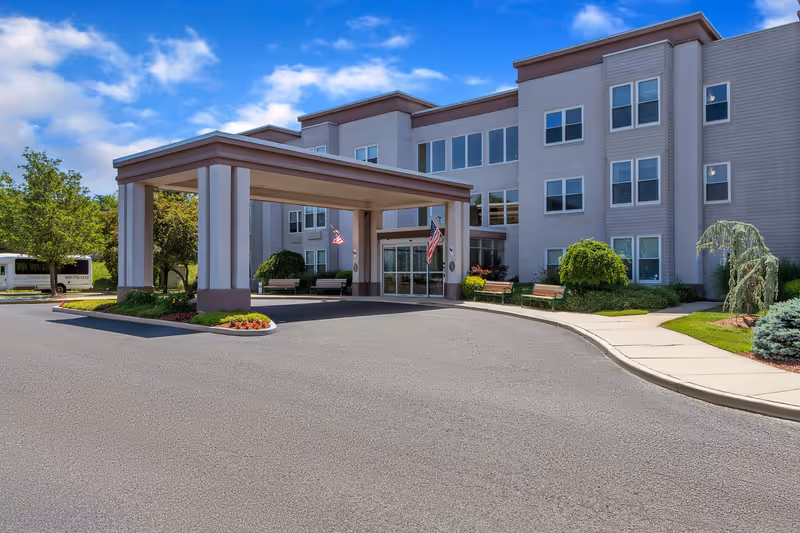 Exterior view of Seaton Voorhees senior living facility showing the main entrance with a covered drop-off area, benches, landscaping with bushes and trees, and a clear blue sky with some clouds.